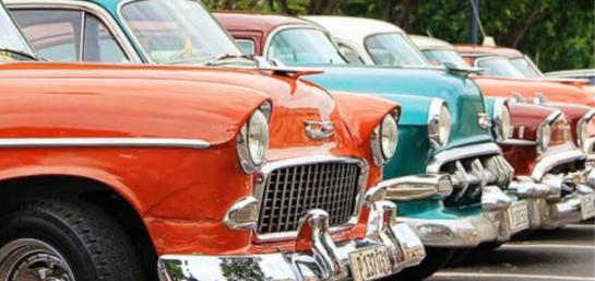 Esta imagen es un primer plano de varios coches antiguos estacionados en fila. El coche más cercano es de color naranja, seguido por uno azul turquesa y otro rojo oscuro. Todos tienen parachoques y parrillas cromadas.