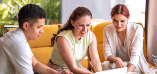 articulo Tres compañeros de trabajo, dos mujeres y un hombre, sentados en un sofá amarillo mirando sonrientes la pantalla de una computadora portátil.